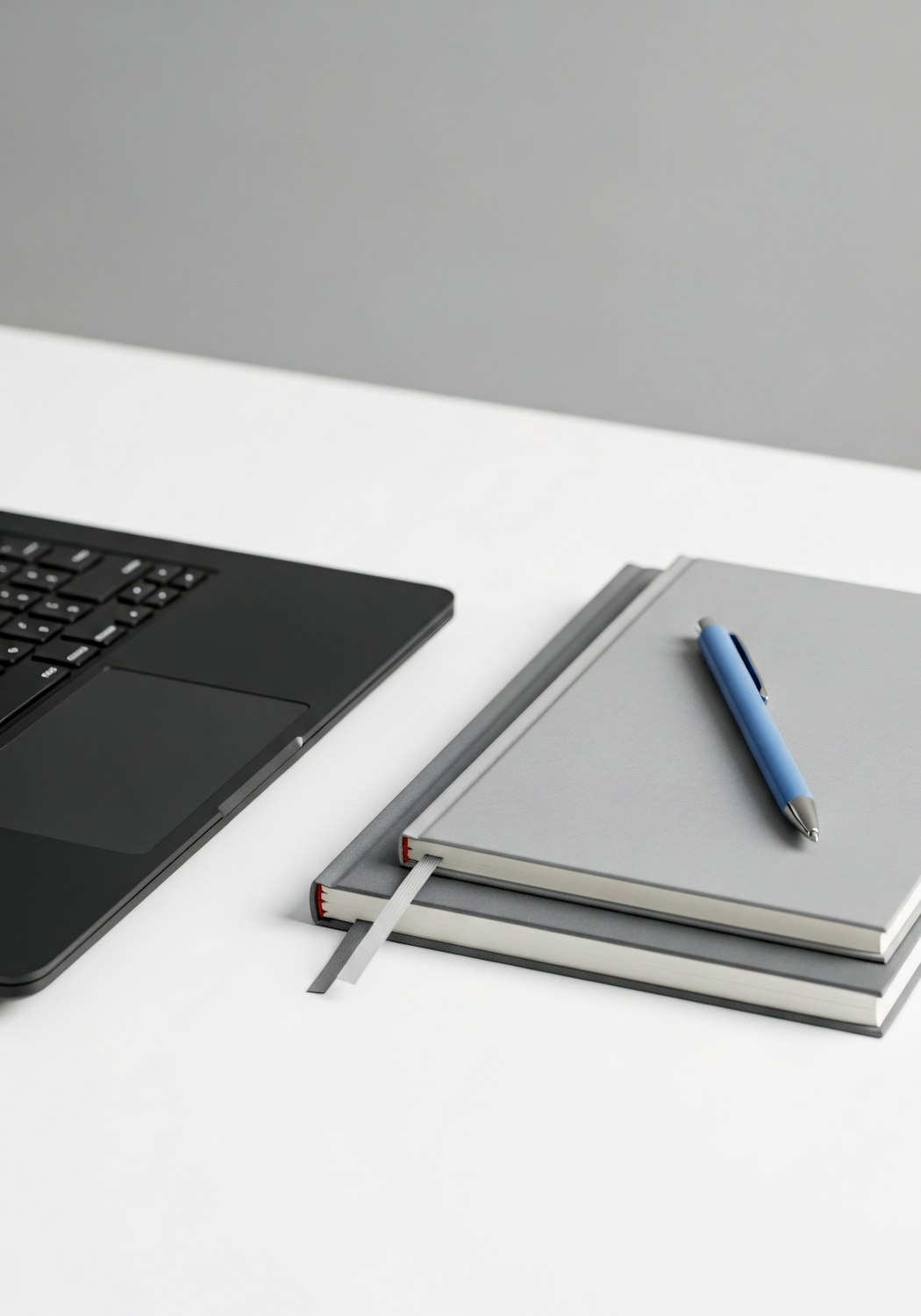 A minimal white desk with a black laptop, two gray notebooks and a blue pen